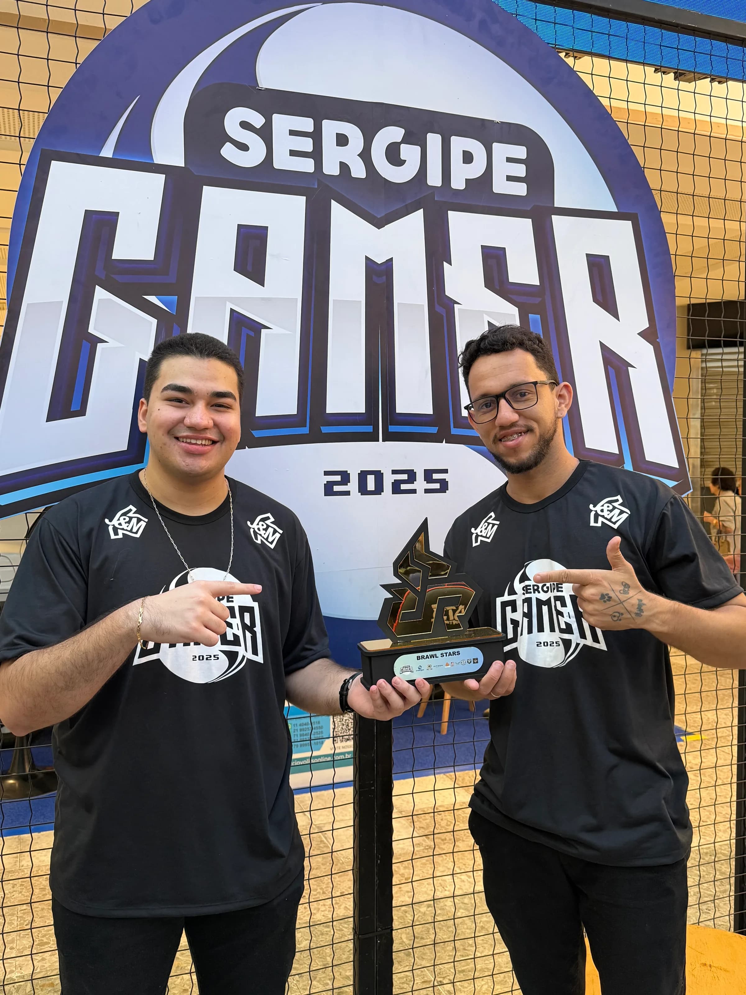 Dupla da Arretados sorrindo e apontando pro troféu do Sergipe Gamer 2025.